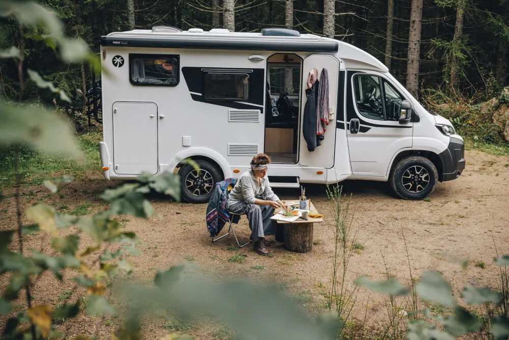 Eine Frau sitzt vor einem Sunlight Camper in einem Wald und isst an einem kleinen Tisch.