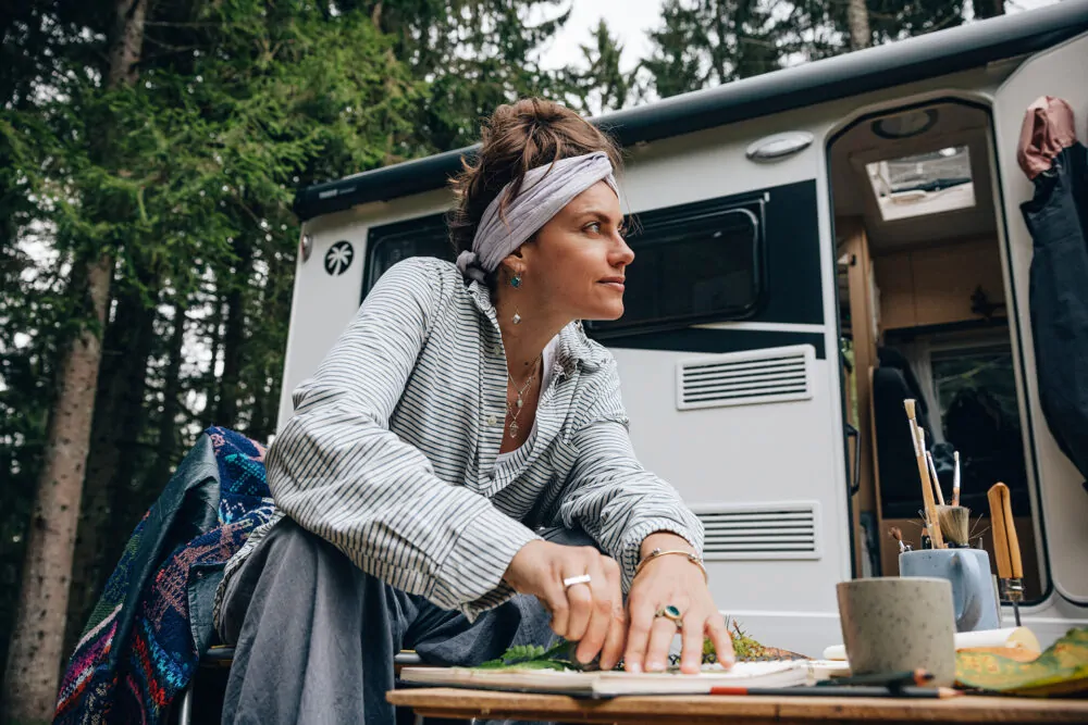 Eine Frau sitzt vor einem Sunlight Van und malt an einem Tisch im Freien.