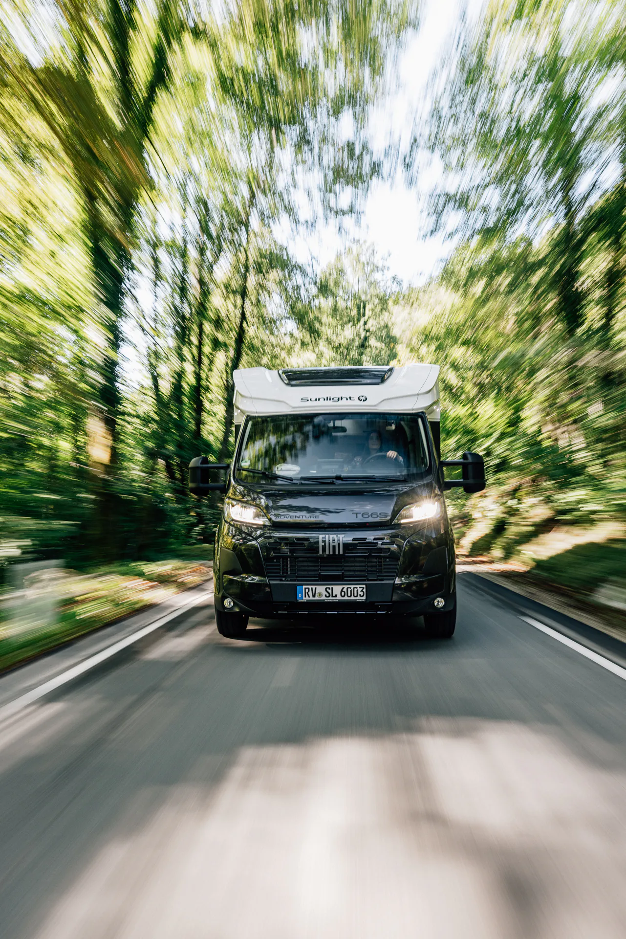 Ein teilintegriertes Wohnmobil der Marke Sunlight Fiat T 669 fährt auf einer Straße durch einen Wald.