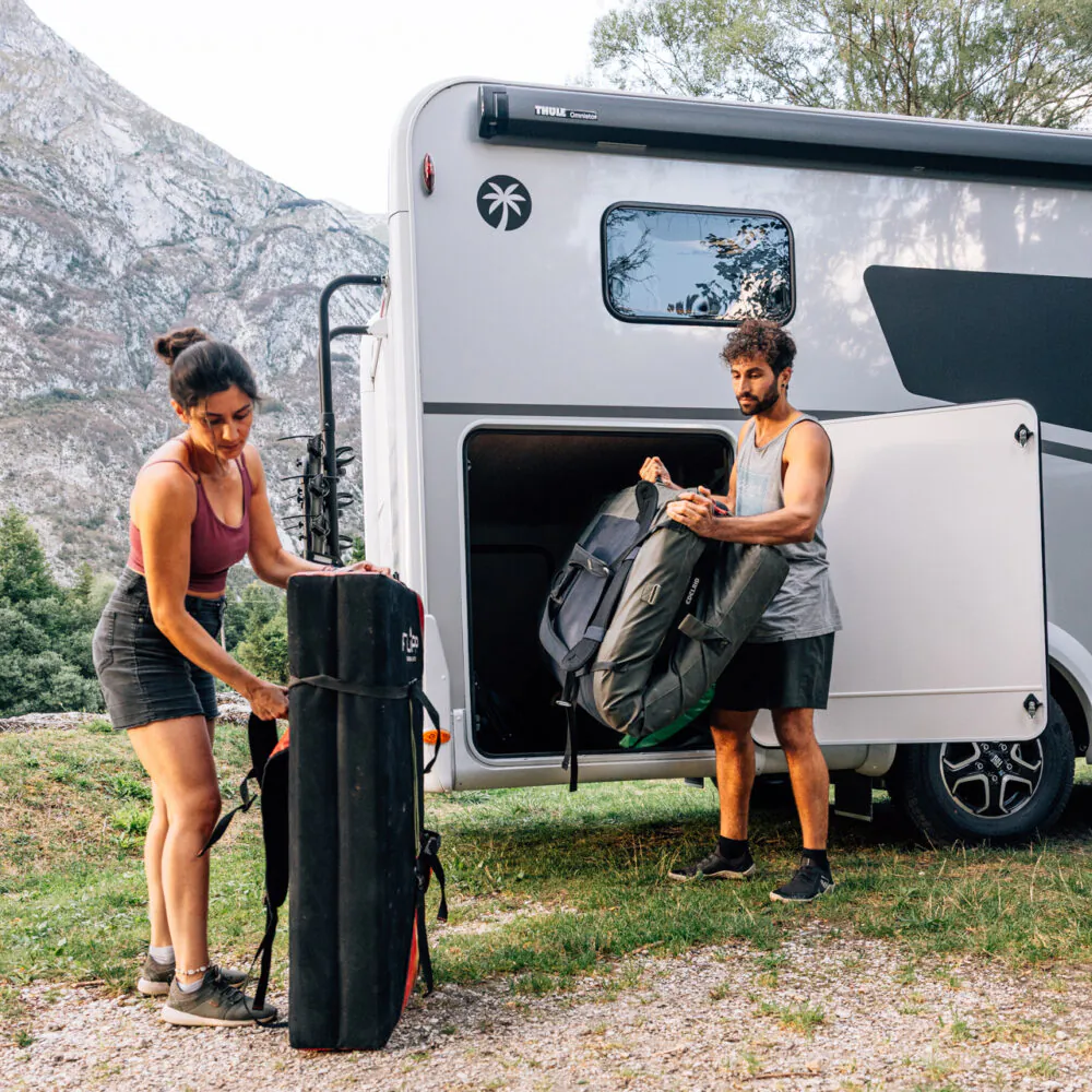Zwei Personen laden Gepäck in ein teilintegriertes Wohnmobil vor einer Bergkulisse.