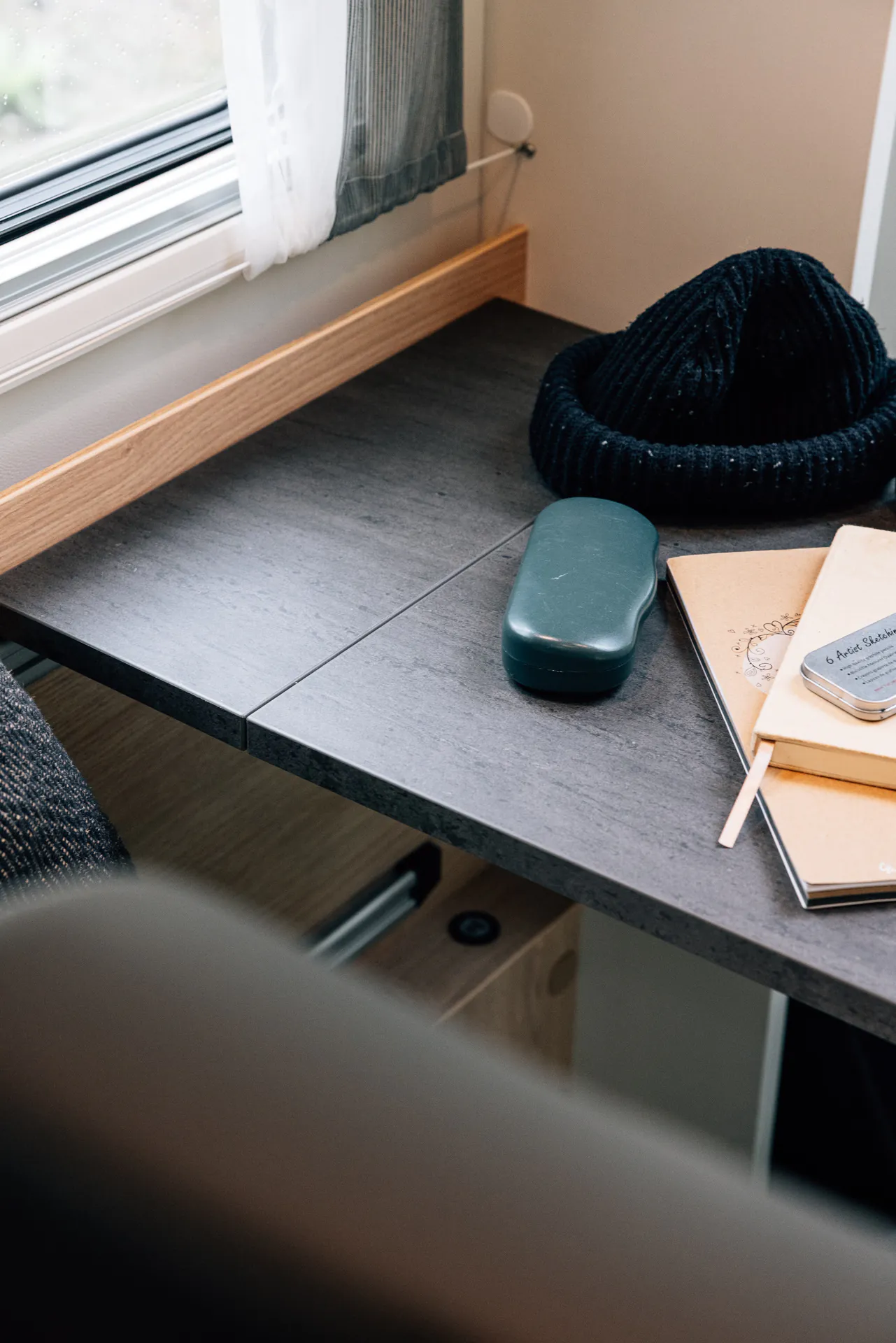 Ein Tisch in einem Wohnmobil mit einem Notizbuch, einer Brille in einem Etui und einer Mütze darauf.