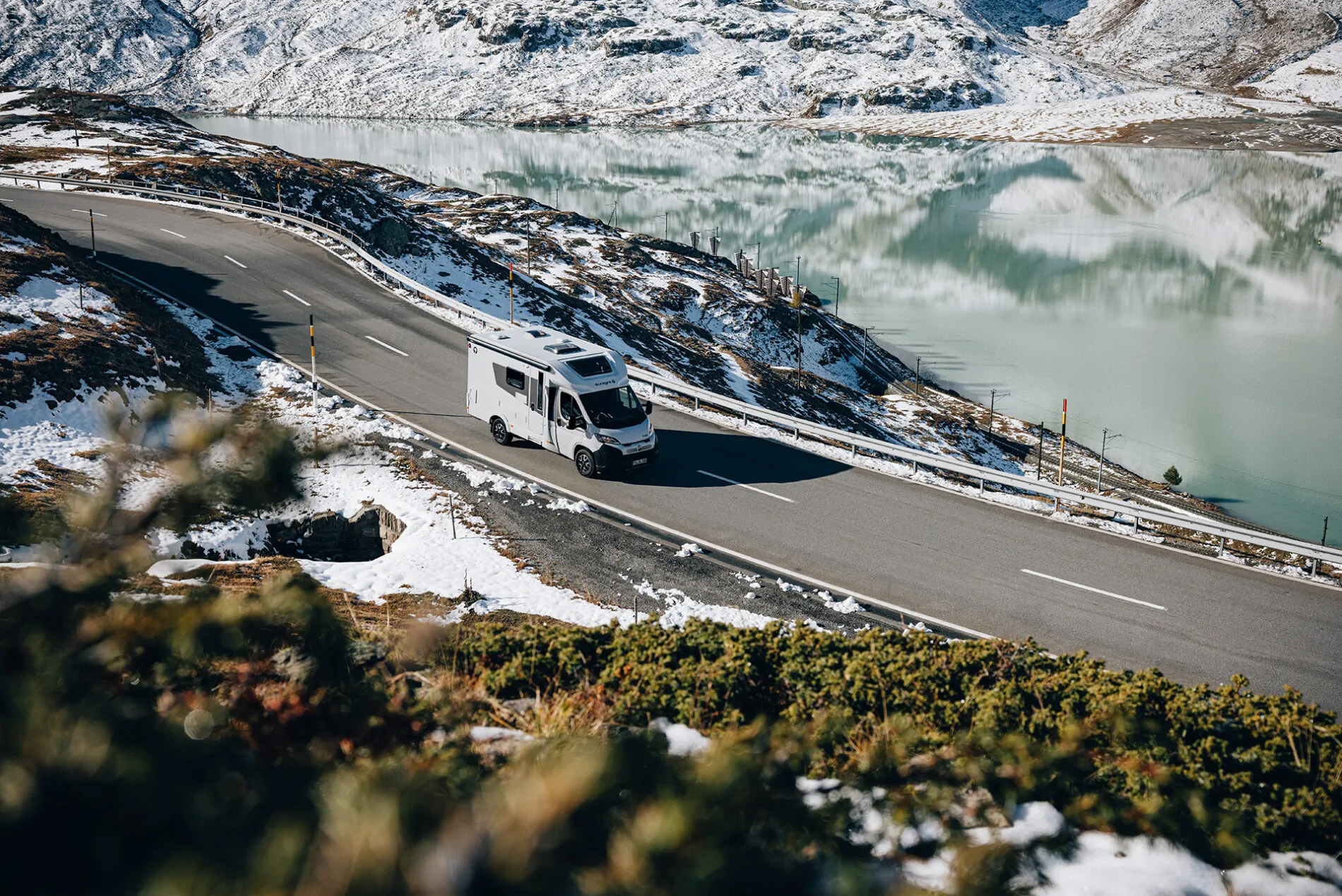 Ein teilintegriertes Wohnmobil fährt auf einer verschneiten Bergstraße entlang eines Sees.