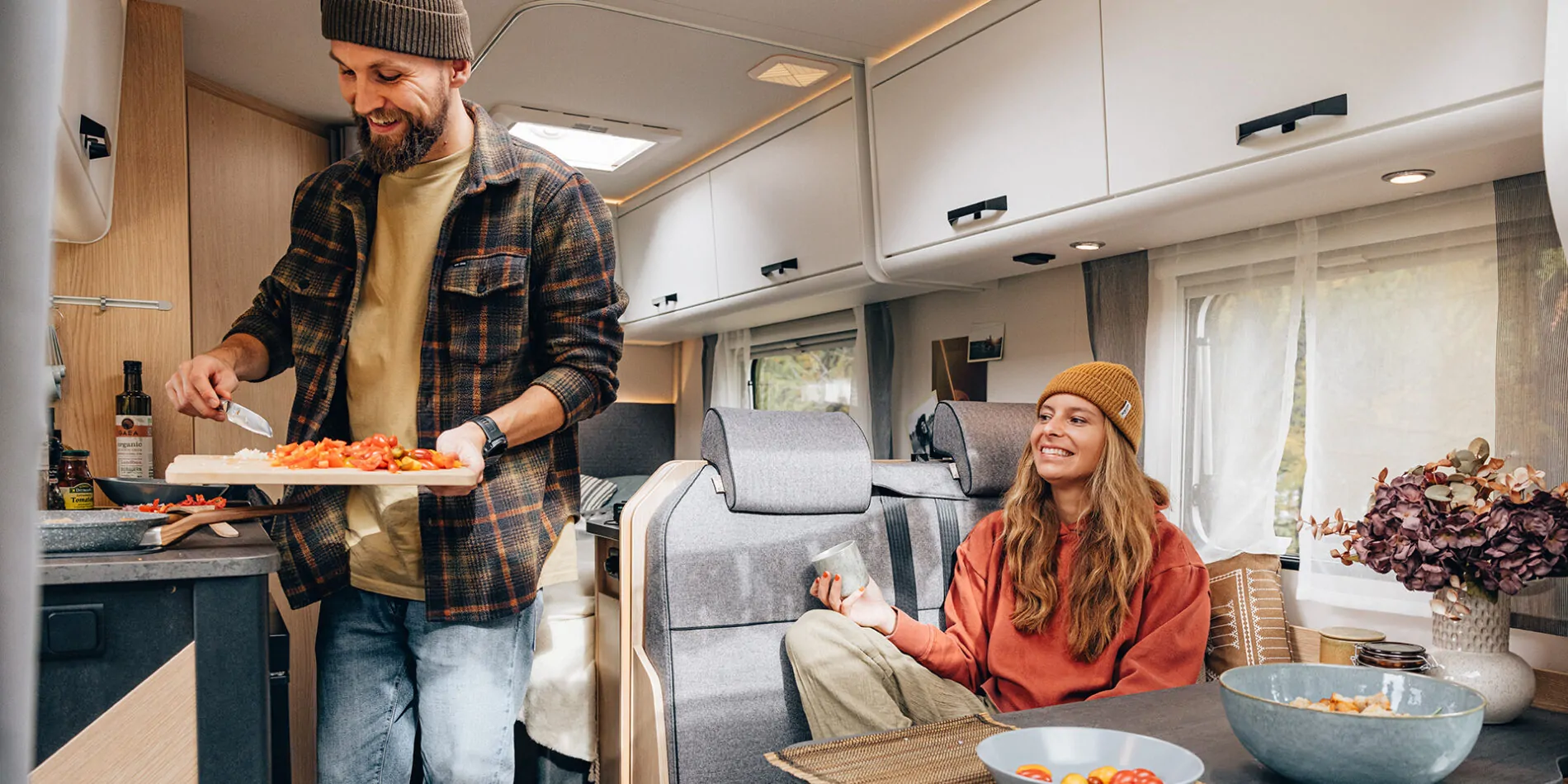 Ein Mann bereitet Essen in der Küche eines teilintegrierten Wohnmobils zu, während eine Frau entspannt auf der Sitzbank sitzt.