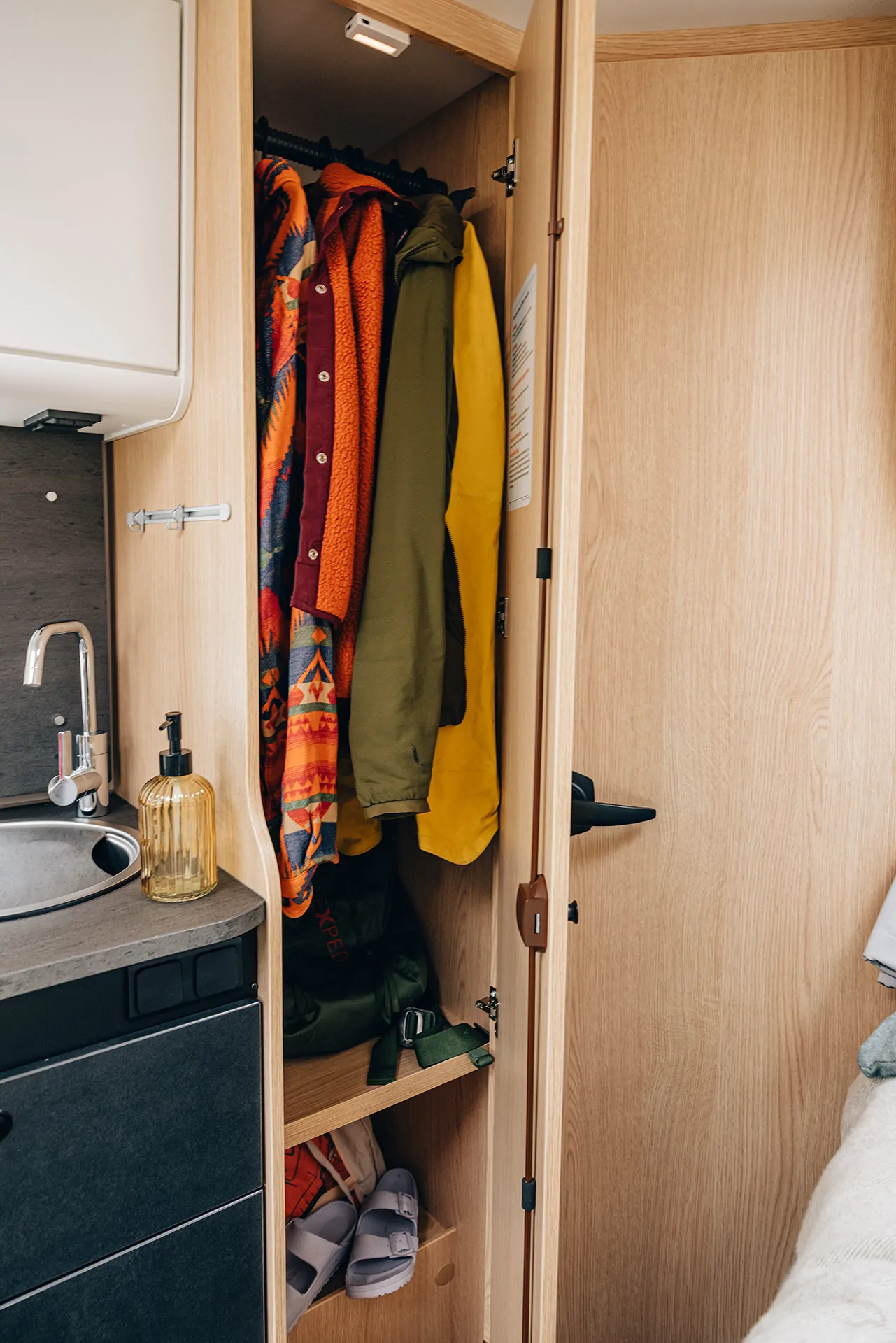 Offener Kleiderschrank in einem Wohnmobil mit bunten Jacken und einem Regal mit Hausschuhen.