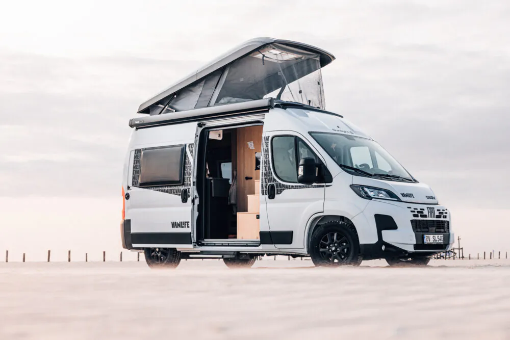 Weißer Campervan mit geöffnetem Aufstelldach und Schiebetür in einer sandigen Landschaft.