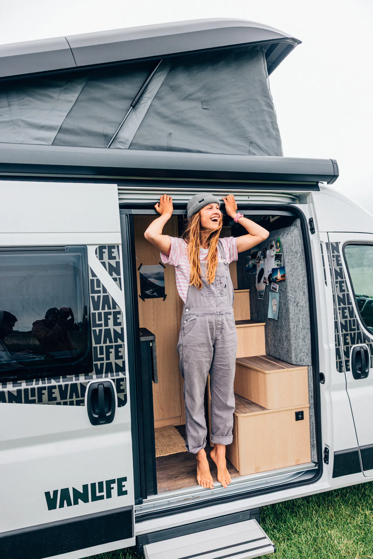 Eine Person steht lächelnd barfuß im Eingang eines Wohnmobils mit der Aufschrift "Vanlife".