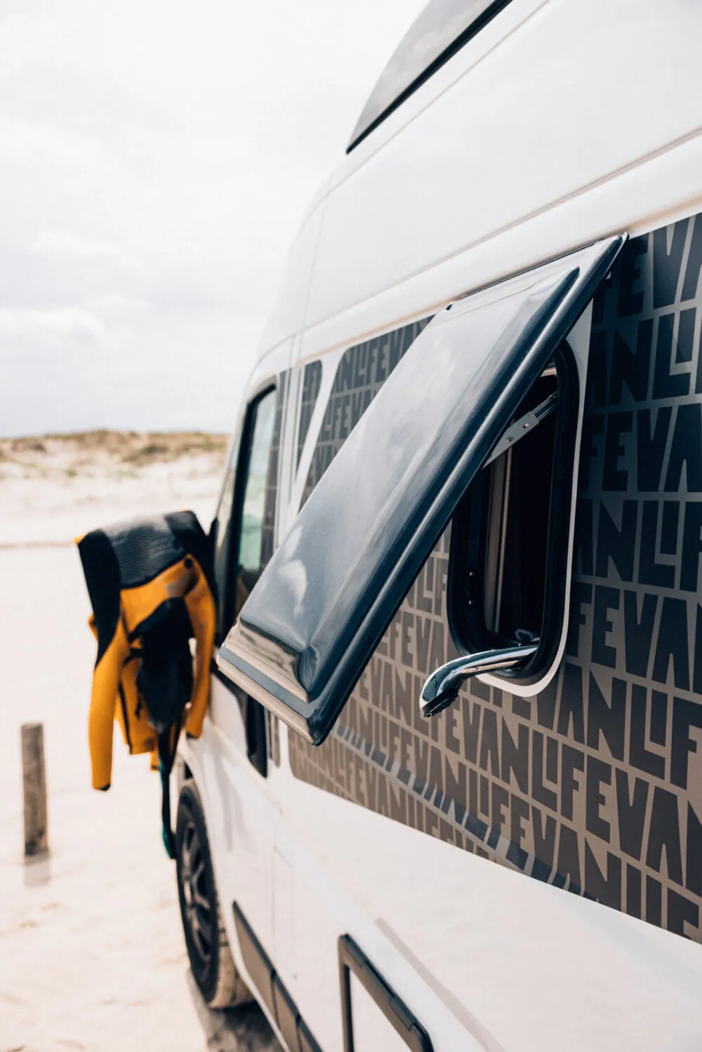 Seitenansicht eines Campervans mit geöffnetem Fenster und aufgehängtem Neoprenanzug an einem Strand.