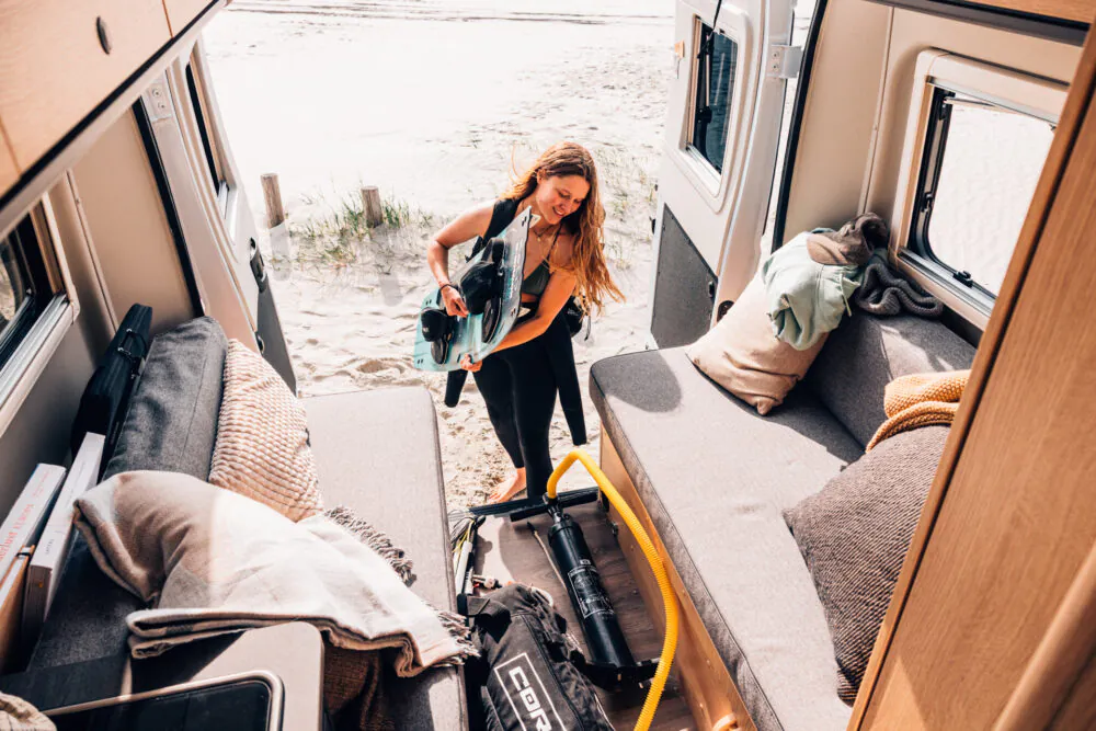 Eine Frau verstaut ein Surfbrett im Interieur eines Campingvans am Strand.