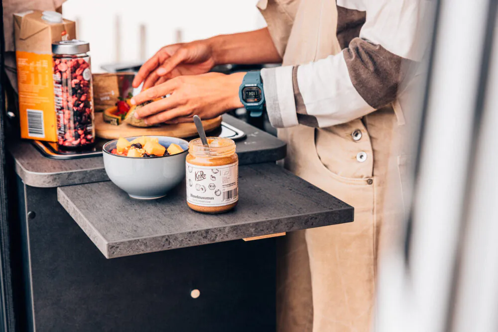 Person bereitet Essen in einer Camper-Küche mit Obst und Nussmus zu.