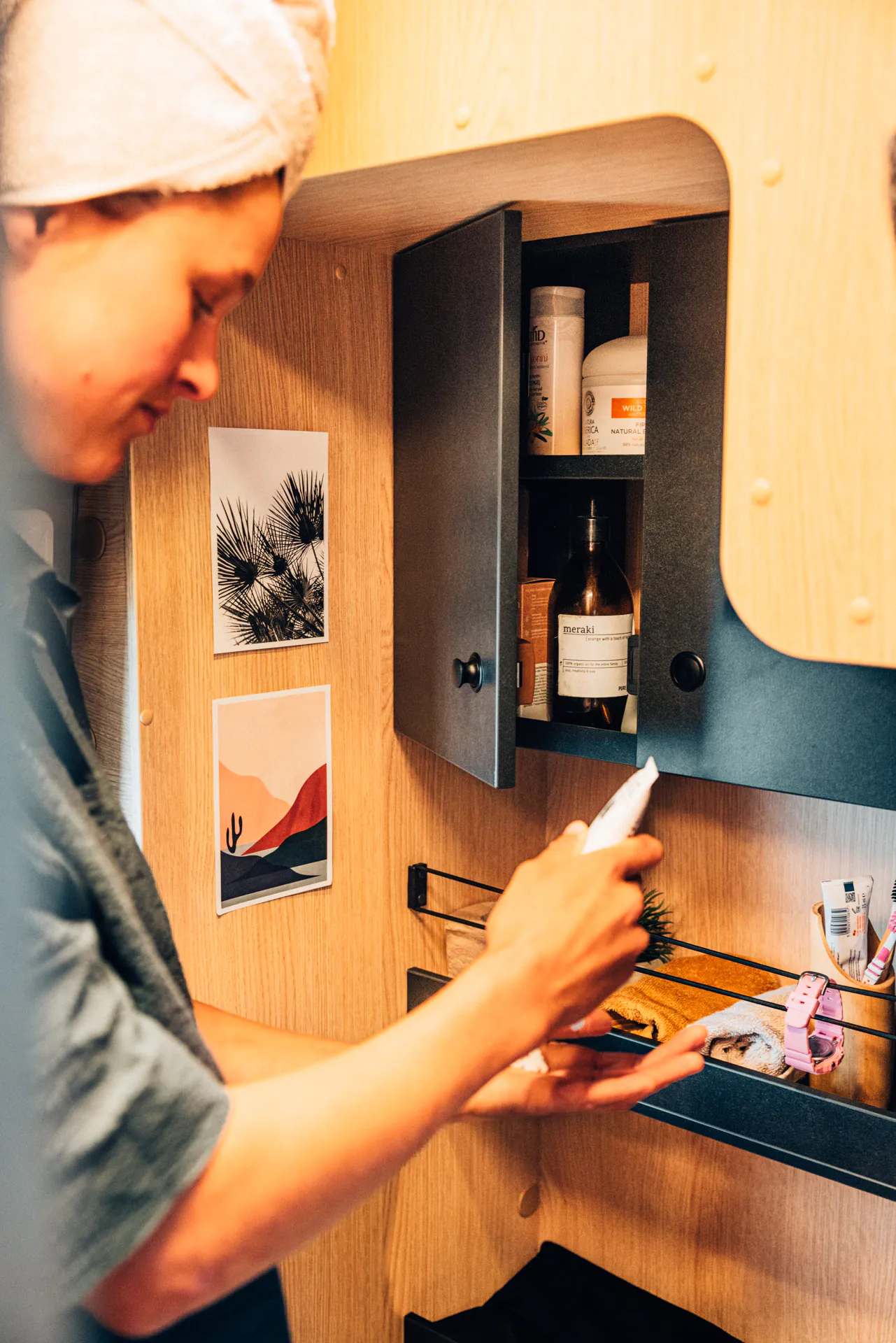 Eine Person trägt ein Handtuch auf dem Kopf und benutzt Hautpflegeprodukte vor einem geöffneten Schrank im Camper.