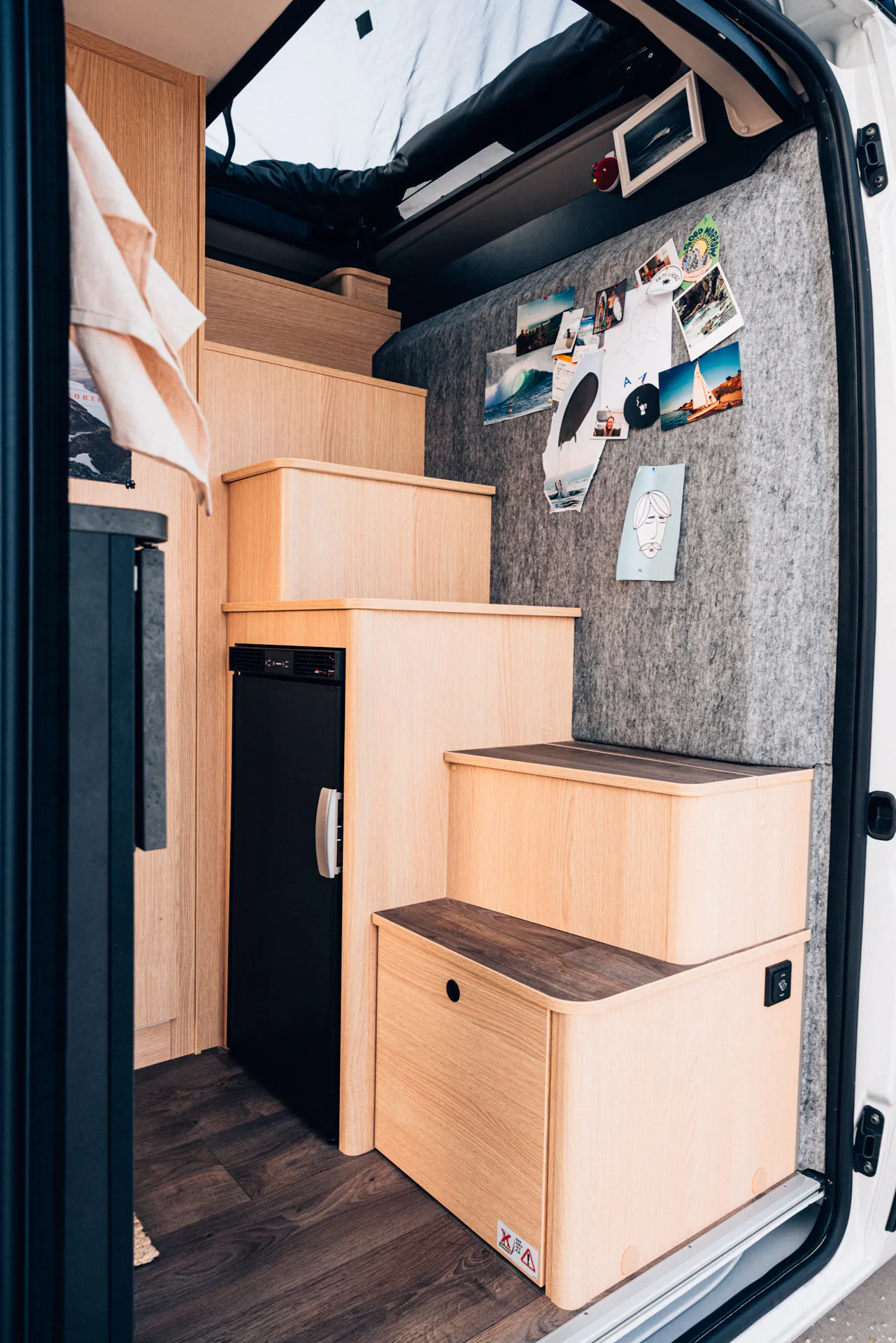 Innenansicht eines Campervans mit Holztreppen, einem kleinen Kühlschrank und an der Wand befestigten Fotos.