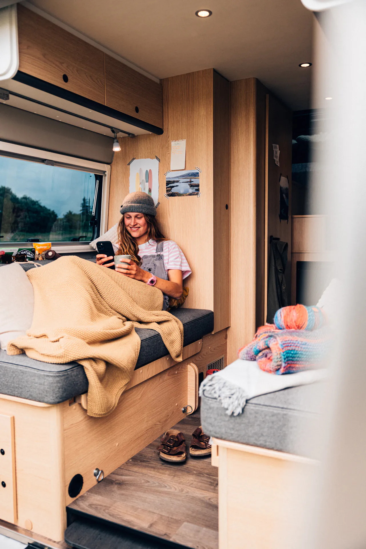 Person entspannt in einem gemütlichen Camper-Interieur mit Handy und Getränk.