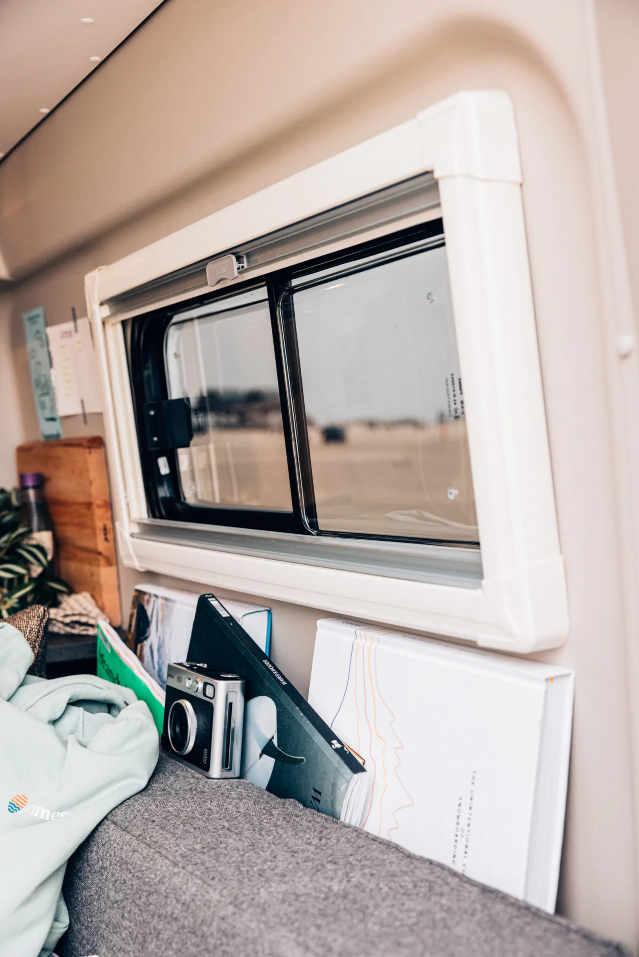 Innenraum eines Wohnmobils mit einem Fenster, einer Kamera und Büchern auf einer grauen Sitzfläche.