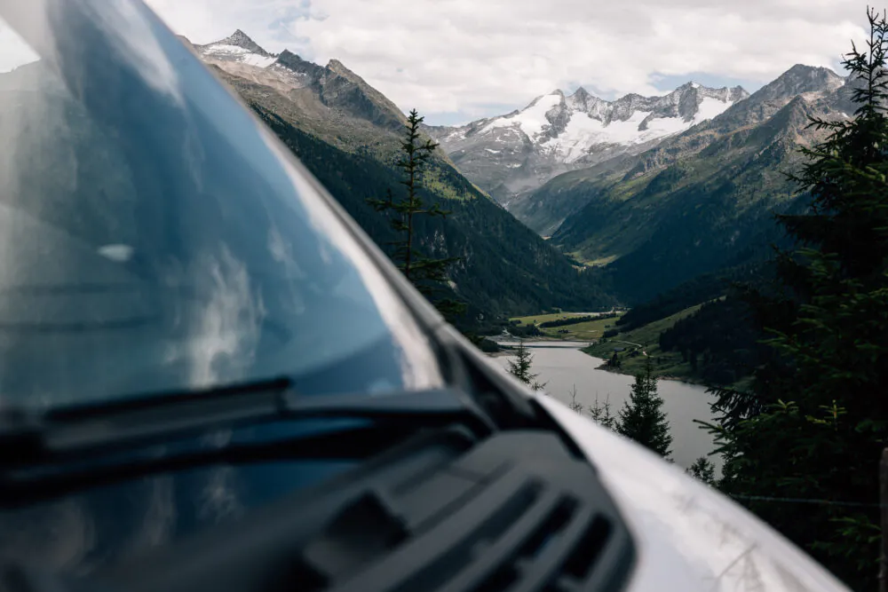Berglandschaft mit See im Tal, im Vordergrund ein Fahrzeug.