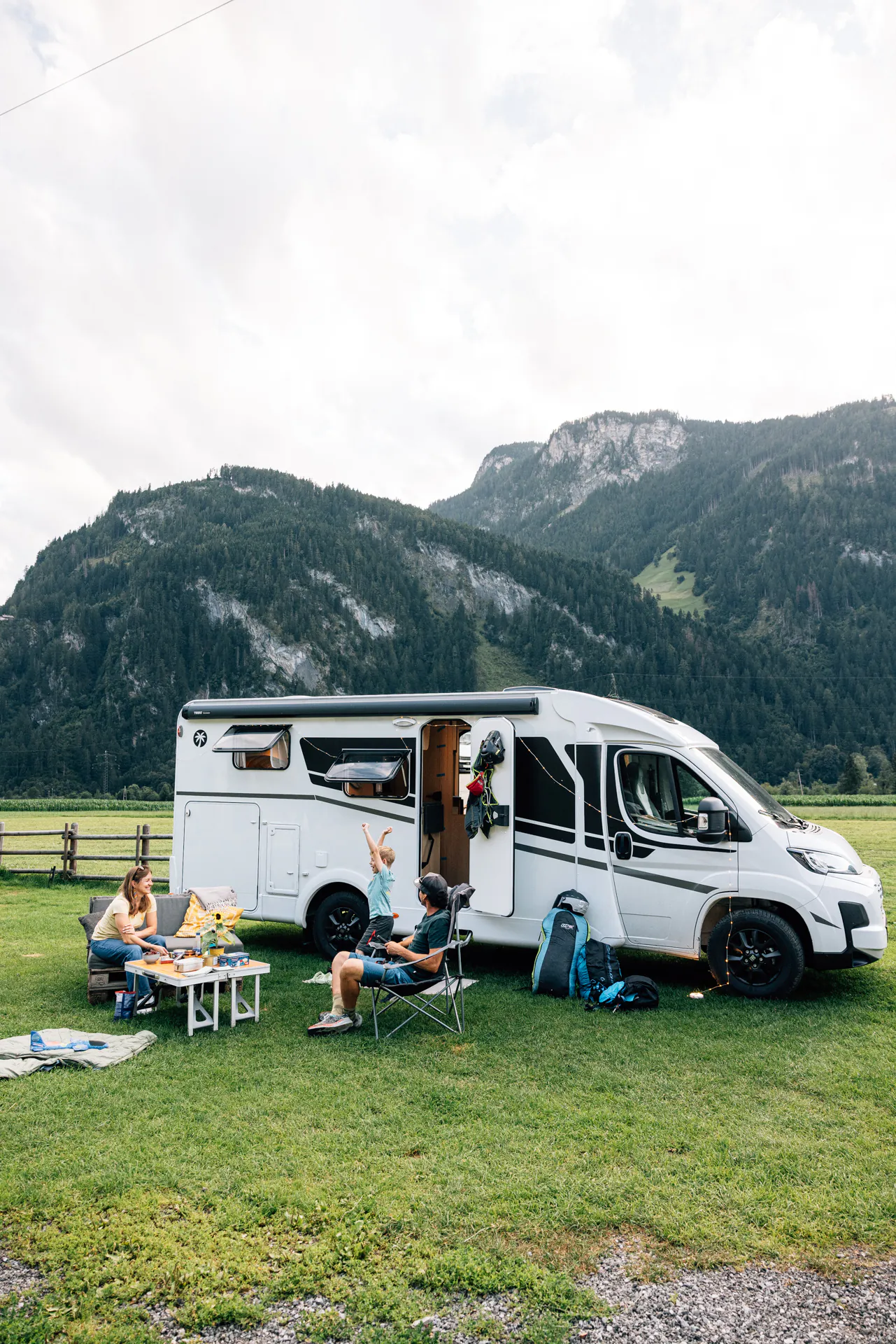 Familie sitzt vor einem weißen Campervan auf einer Wiese mit Bergen im Hintergrund.