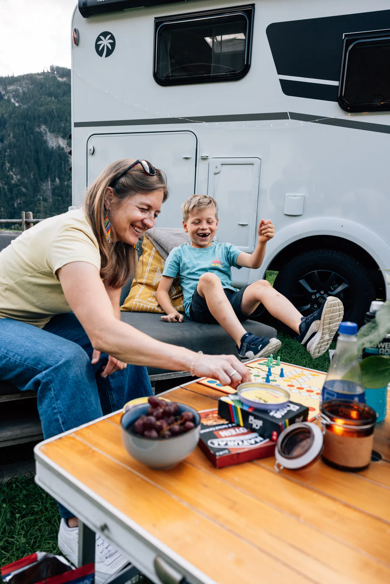 Eine Frau und ein Junge spielen lachend ein Brettspiel neben einem Wohnmobil.