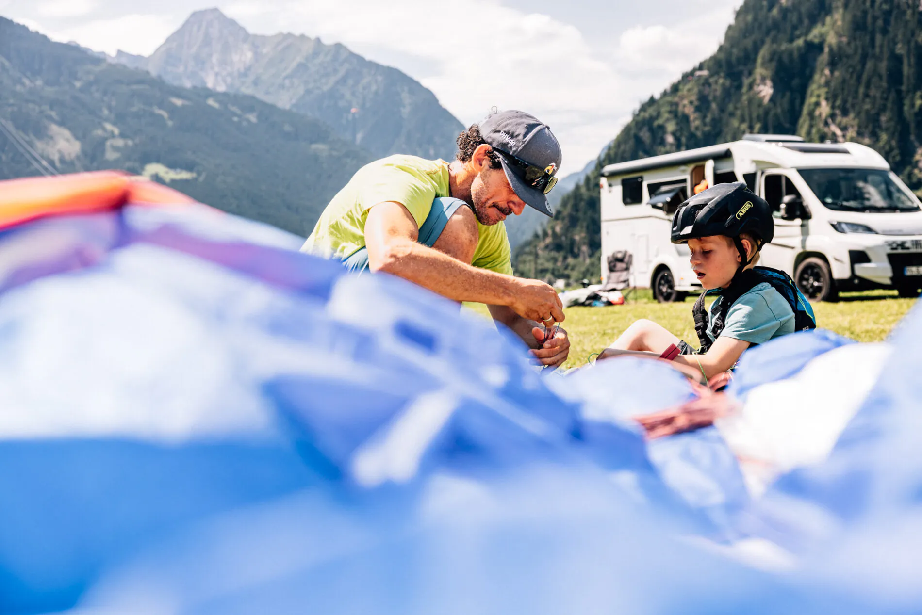 Ein Mann und ein Kind sitzen auf einer Wiese vor einem Wohnmobil, umgeben von Bergen.