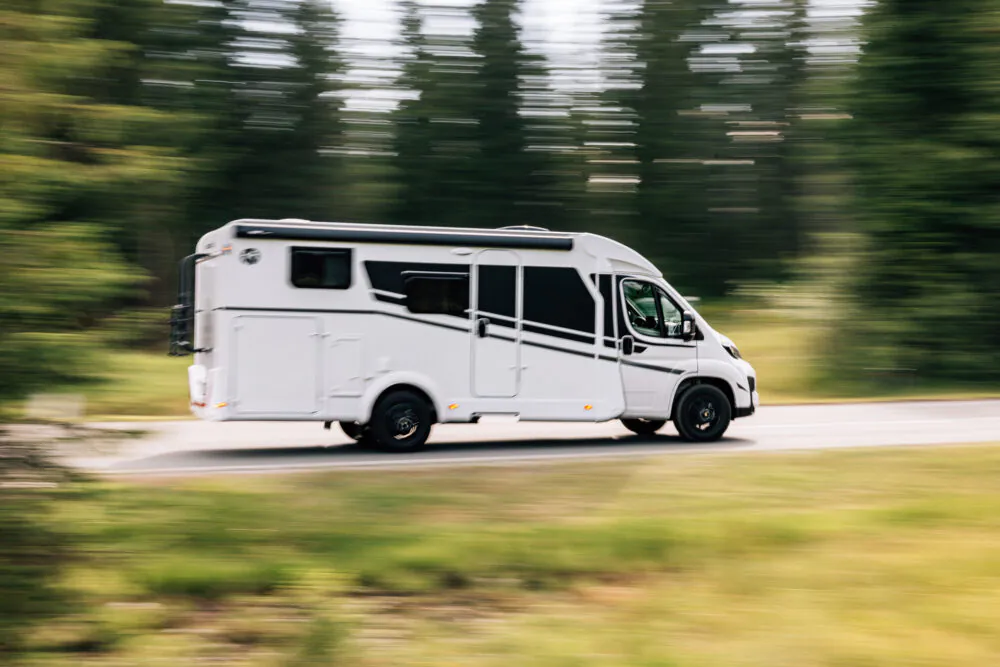 Ein weißer Campervan fährt schnell auf einer Straße durch eine bewaldete Landschaft.