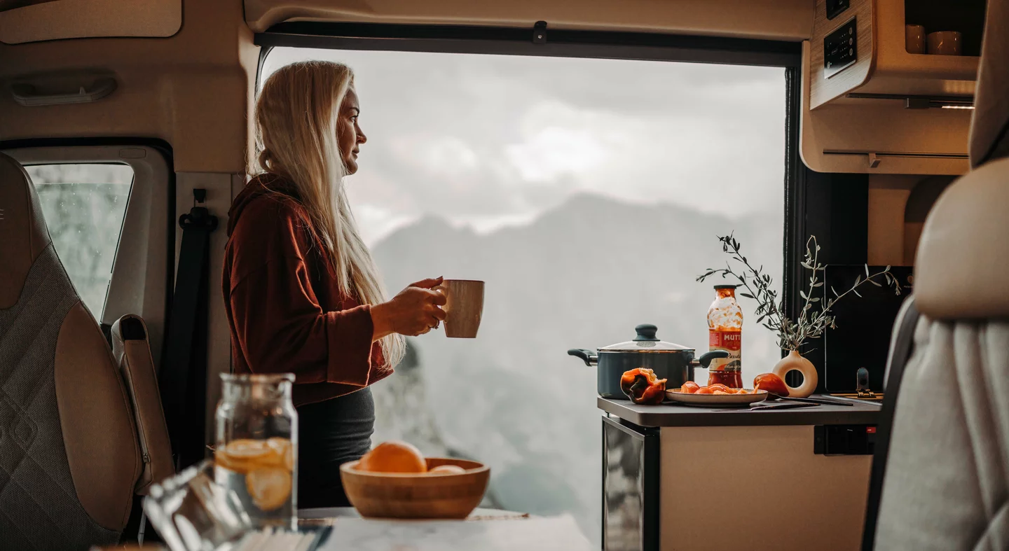 Eine Person steht in einem Camper und hält eine Tasse, während sie aus dem Fenster auf eine Berglandschaft schaut.