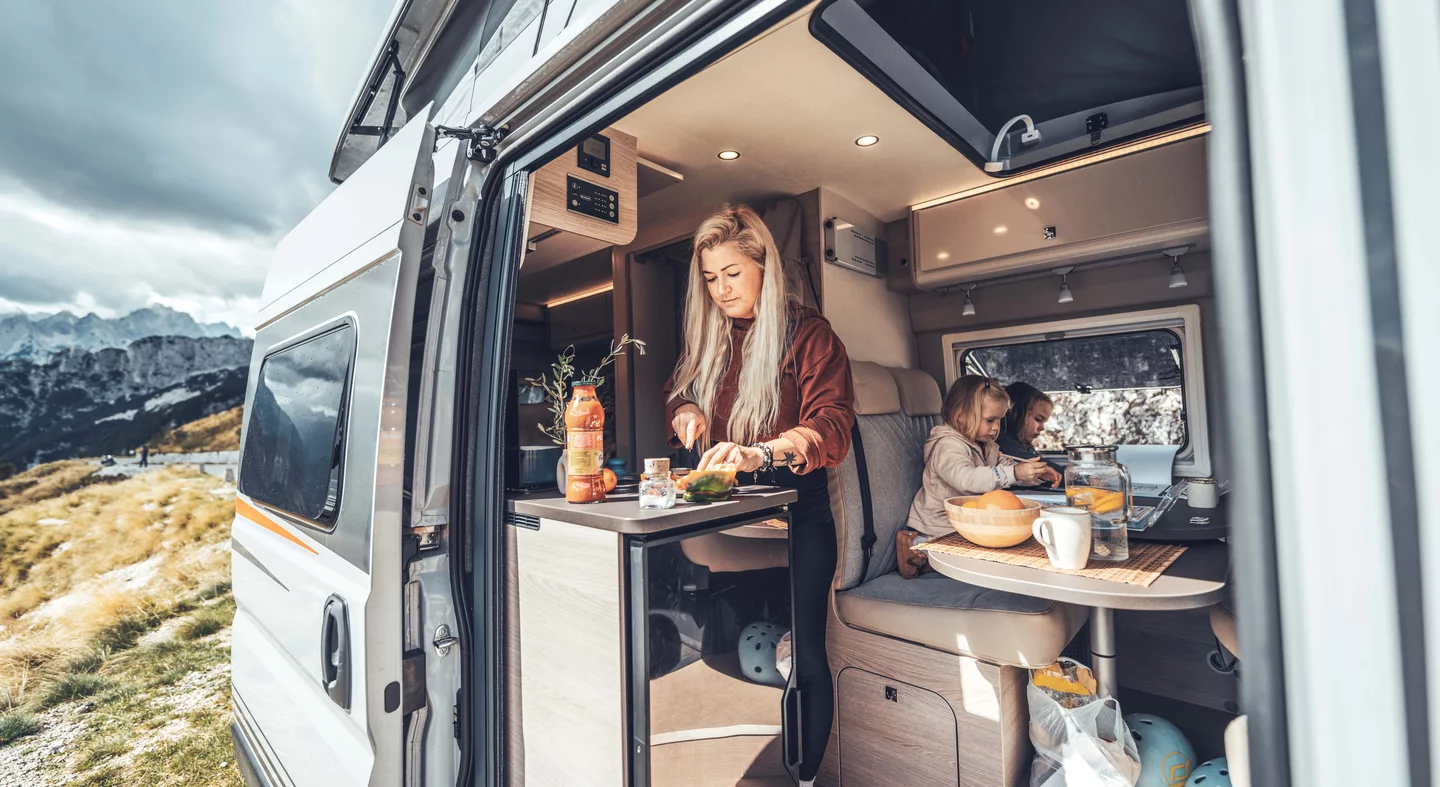 Eine Frau bereitet in einem Campeoc Wohnmobil Essen zu, während zwei Kinder am Tisch sitzen.