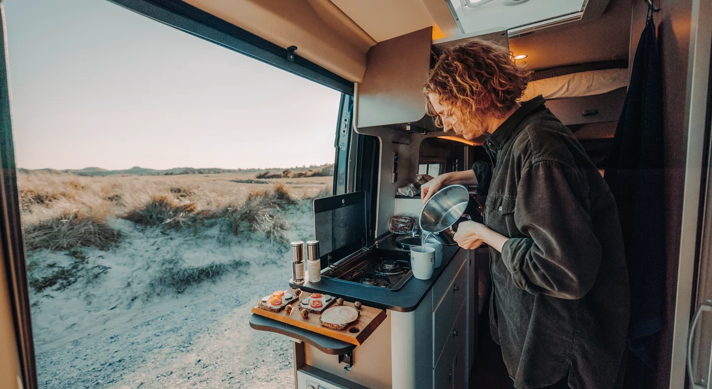 Eine Person kocht in einem Camper, während draußen eine Dünenlandschaft zu sehen ist.