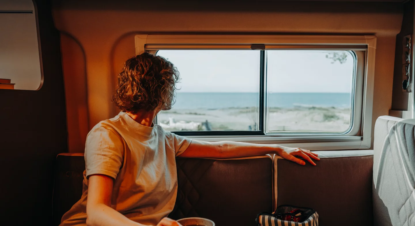 Eine Person sitzt in einem Camper und schaut aus dem Fenster auf das Meer.