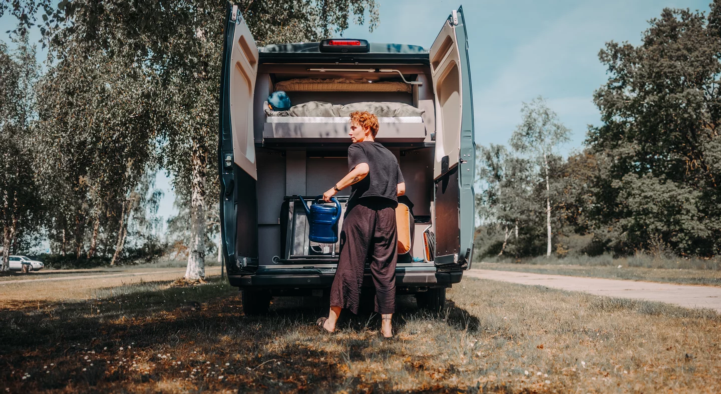 Eine Person steht mit einer Gießkanne am offenen Heck eines Eliseo C644 Campers auf einer Wiese.