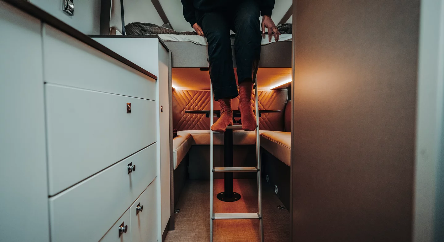 Eine Person sitzt auf einem Hochbett in einem Camper, der durch eine Leiter zugänglich ist.