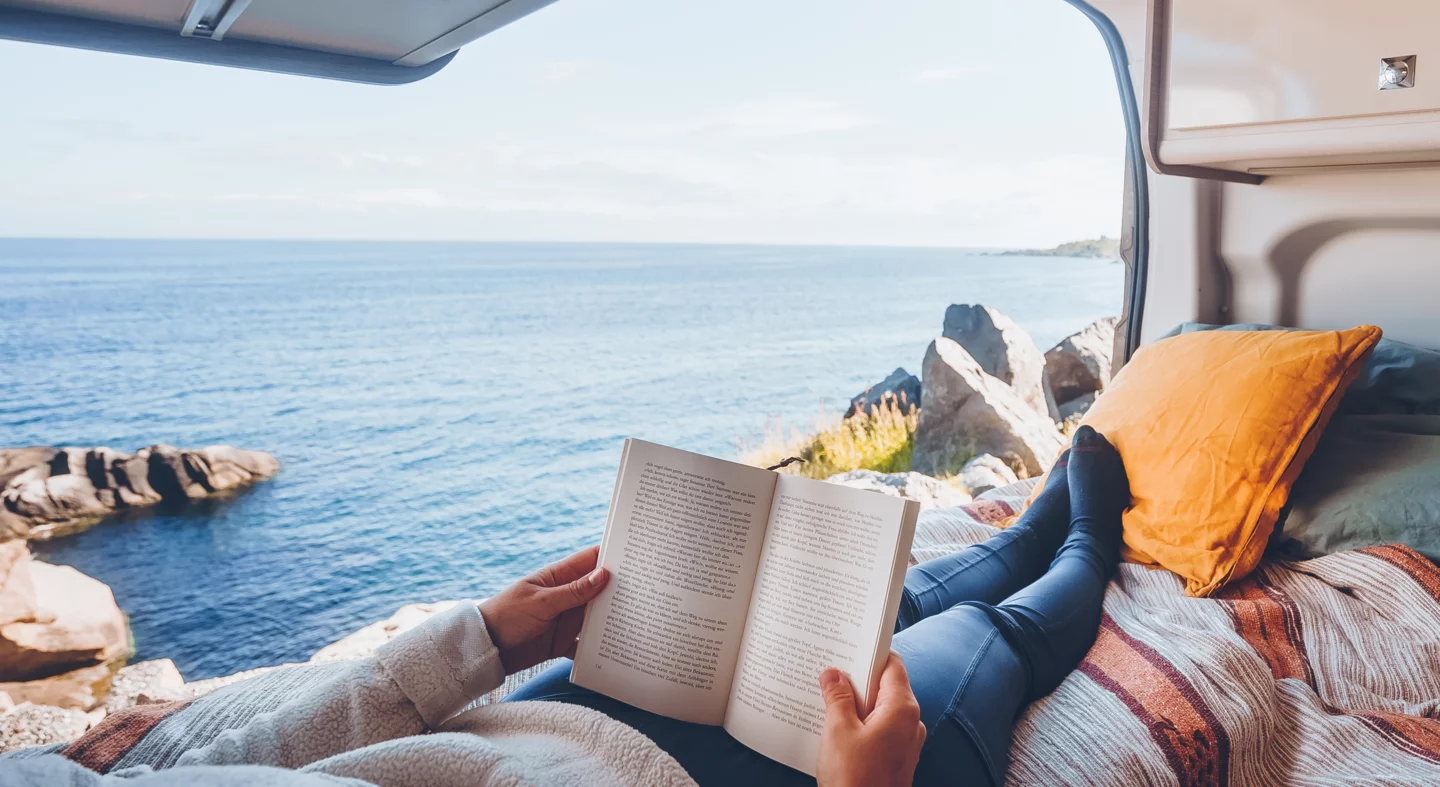Person liest ein Buch in einem Camper mit Blick auf das Meer.