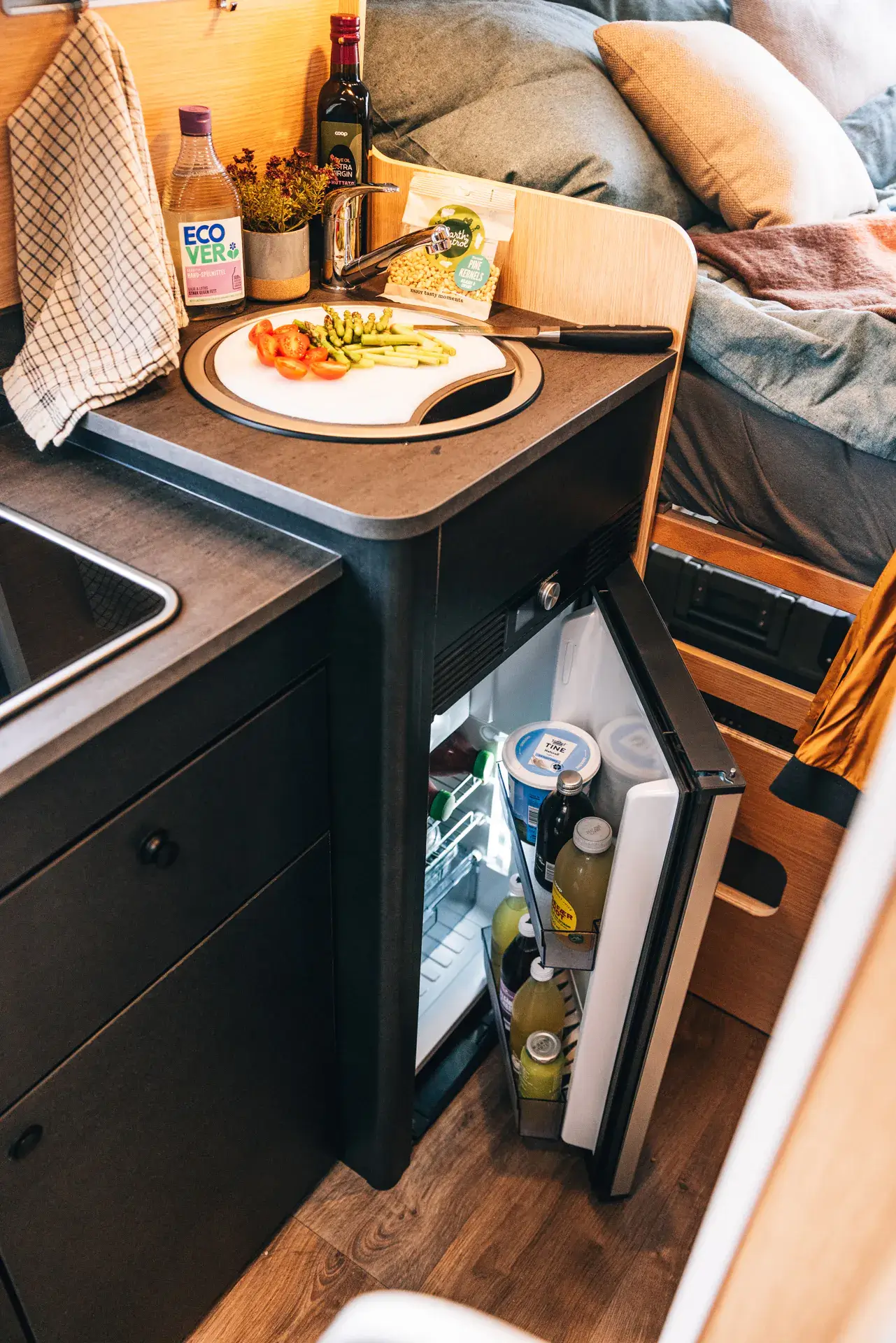 Kleine Küchenzeile im Wohnmobil mit offenem Kühlschrank und Gemüse auf dem Schneidebrett.